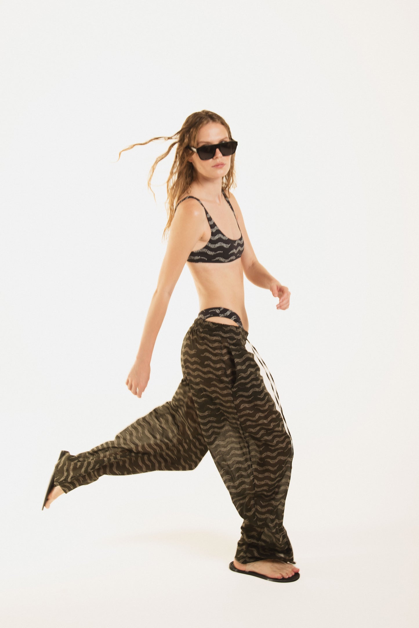 Side view of a girl wearing Nicasia black muslin cotton lightweighted pants with an original print wave-inspired. Premium fabrics, oversized fit. The girl wears also Zoe black bikini 