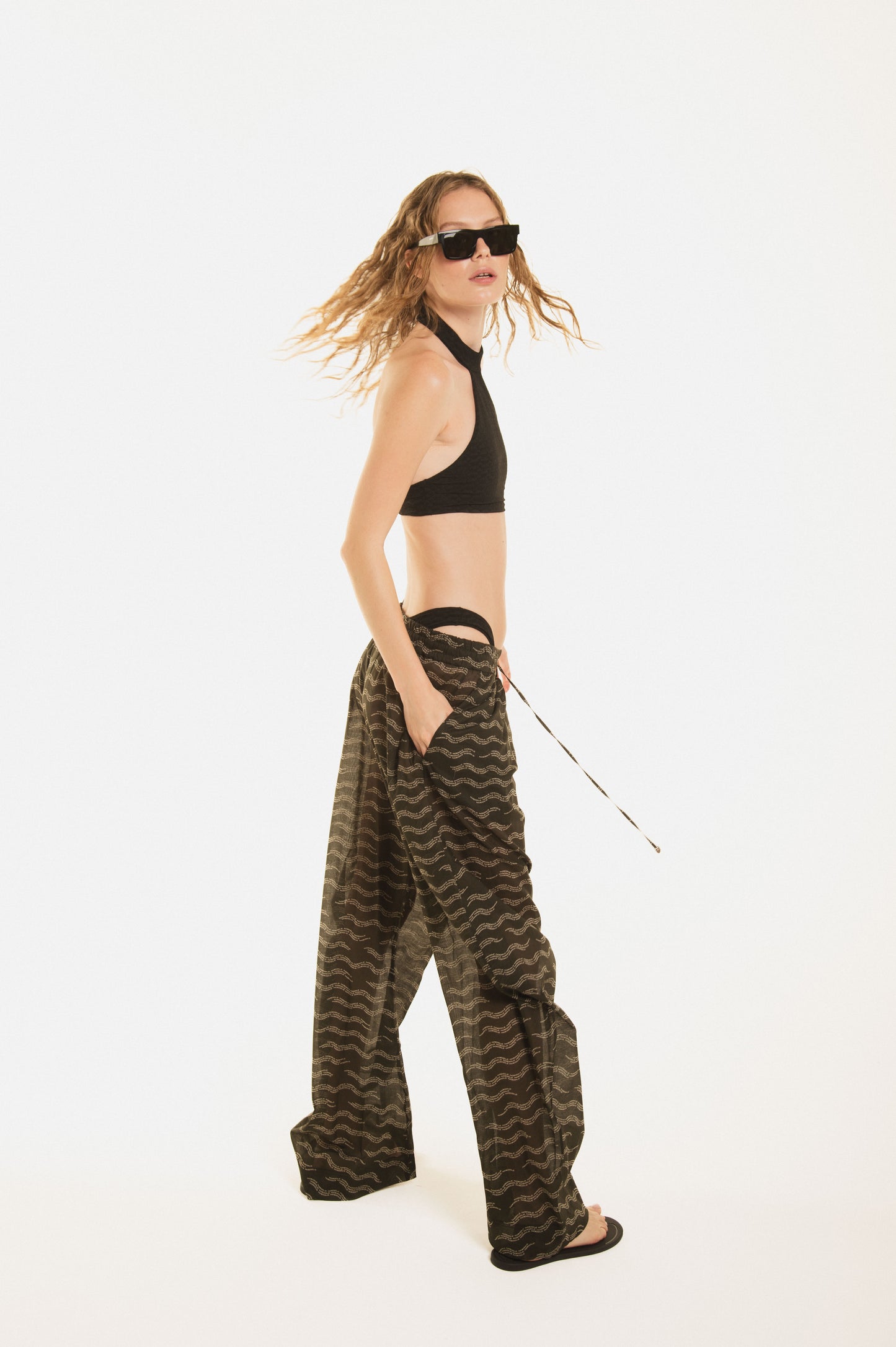
                  
                    Back view of a girl wearing Nicasia black muslin cotton lightweighted pants with an original print wave-inspired. Premium fabrics, oversized fit. The girl wears also Cambon black bikini 
                  
                