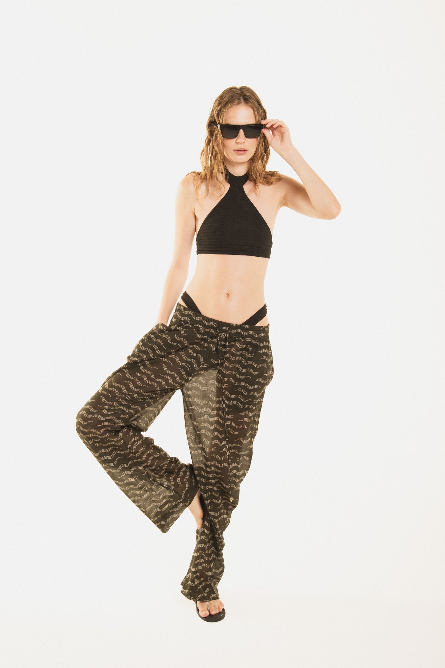 
                  
                    Front view of a girl wearing Nicasia black muslin cotton lightweighted pants with an original print wave-inspired. Premium fabrics, oversized fit. The girl wears also Cambon black bikini 
                  
                