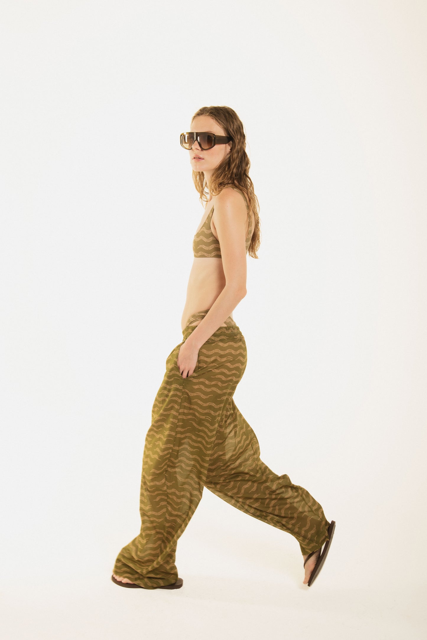 
                  
                    side view of girl walking and wearing Noodle Beachwear wide-leg trousers in fluid fabric with an exclusive olive green wave print. Low-rise waist with drawstring tie, relaxed fit and effortless drape. From beach to dinner. Made in Italy.
                  
                
