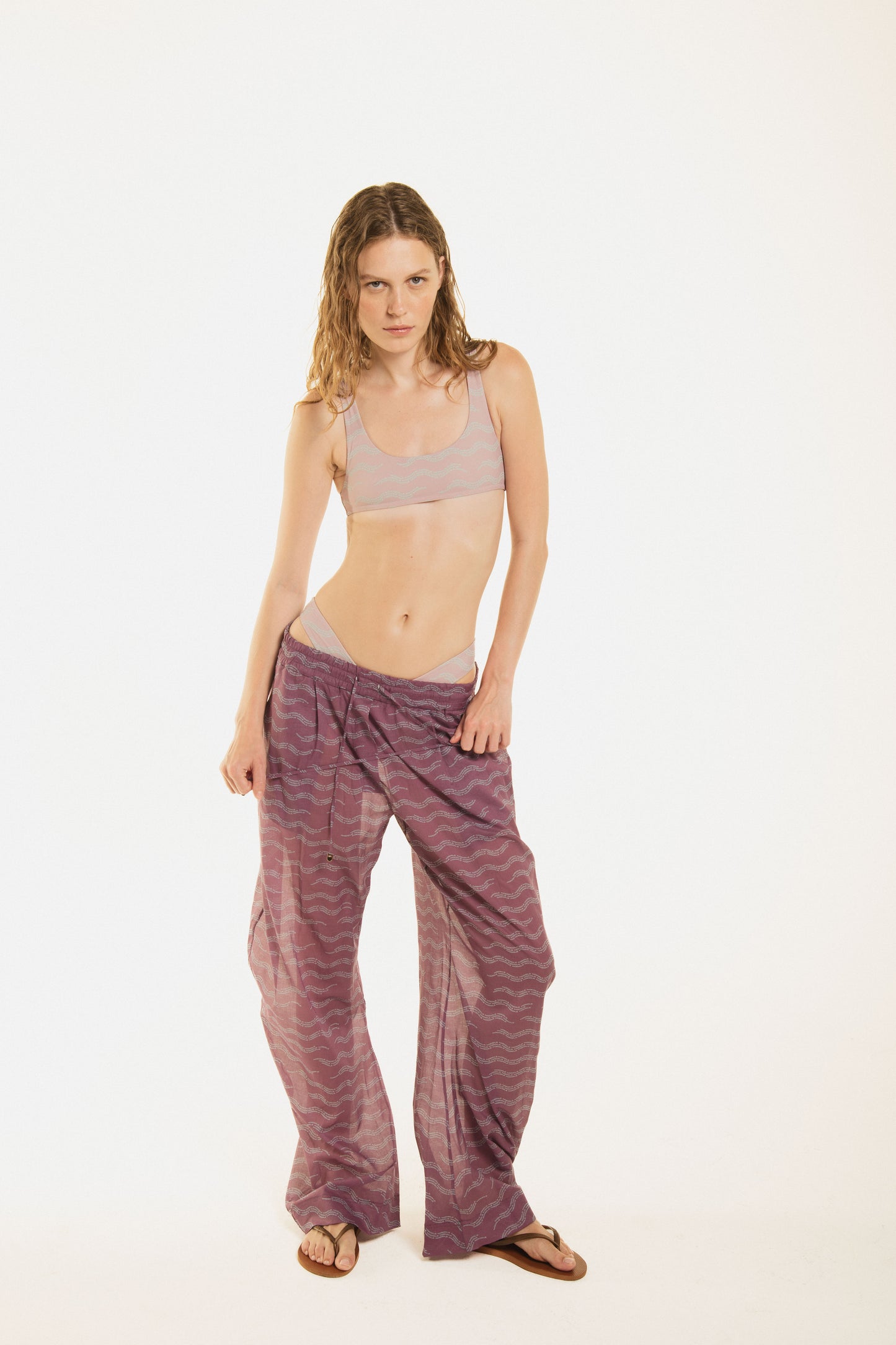 
                  
                    Girl wearing Nicasia grape violet muslin cotton lightweighted pants with an original print wave-inspired. Premium fabrics, oversized fit. The girl wears also Zoe pink bikini 
                  
                