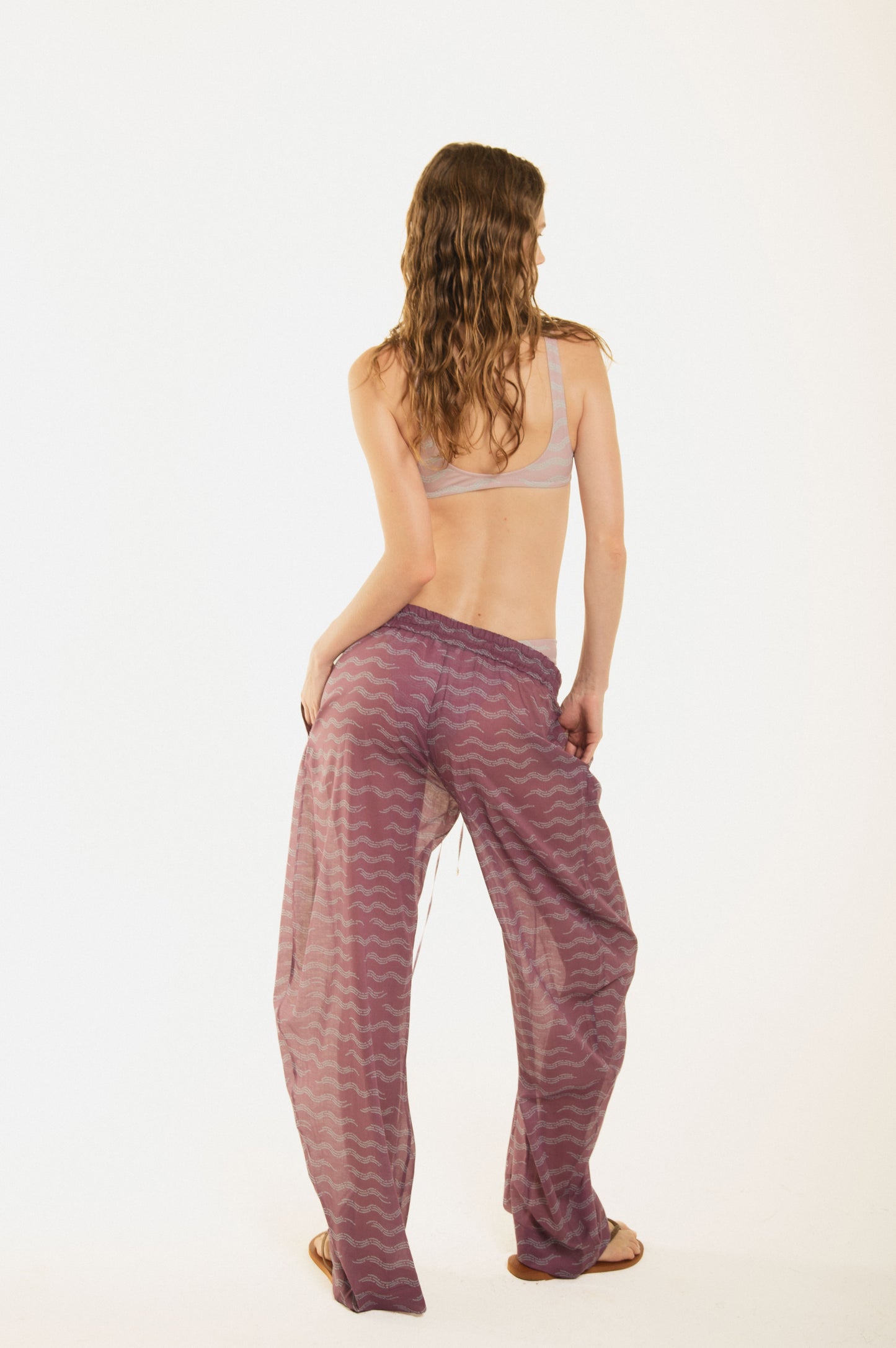 Back view of a girl wearing Nicasia grape violet muslin cotton lightweighted pants with an original print wave-inspired. Premium fabrics, oversized fit. The girl wears also Zoe pink bikini 
