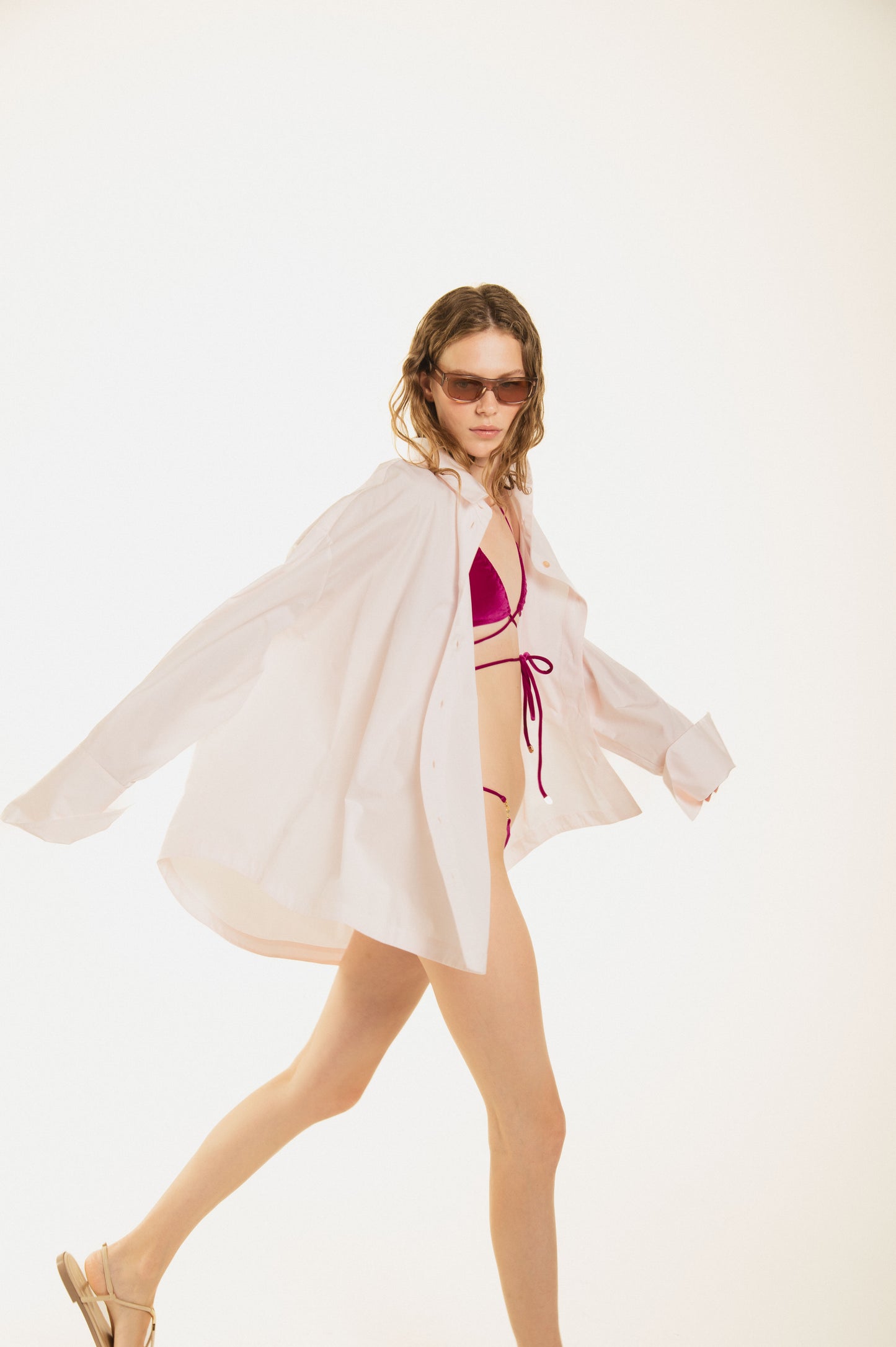 
                  
                    side view of girl walking Noodle Beachwear oversize shirt in blush pink cotton with a curved row of gold jewel buttons. Maxi fit, long sleeves and effortless drape — the ultimate luxury beach cover-up. Made in Italy. The girl wears  tange purple bikini
                  
                