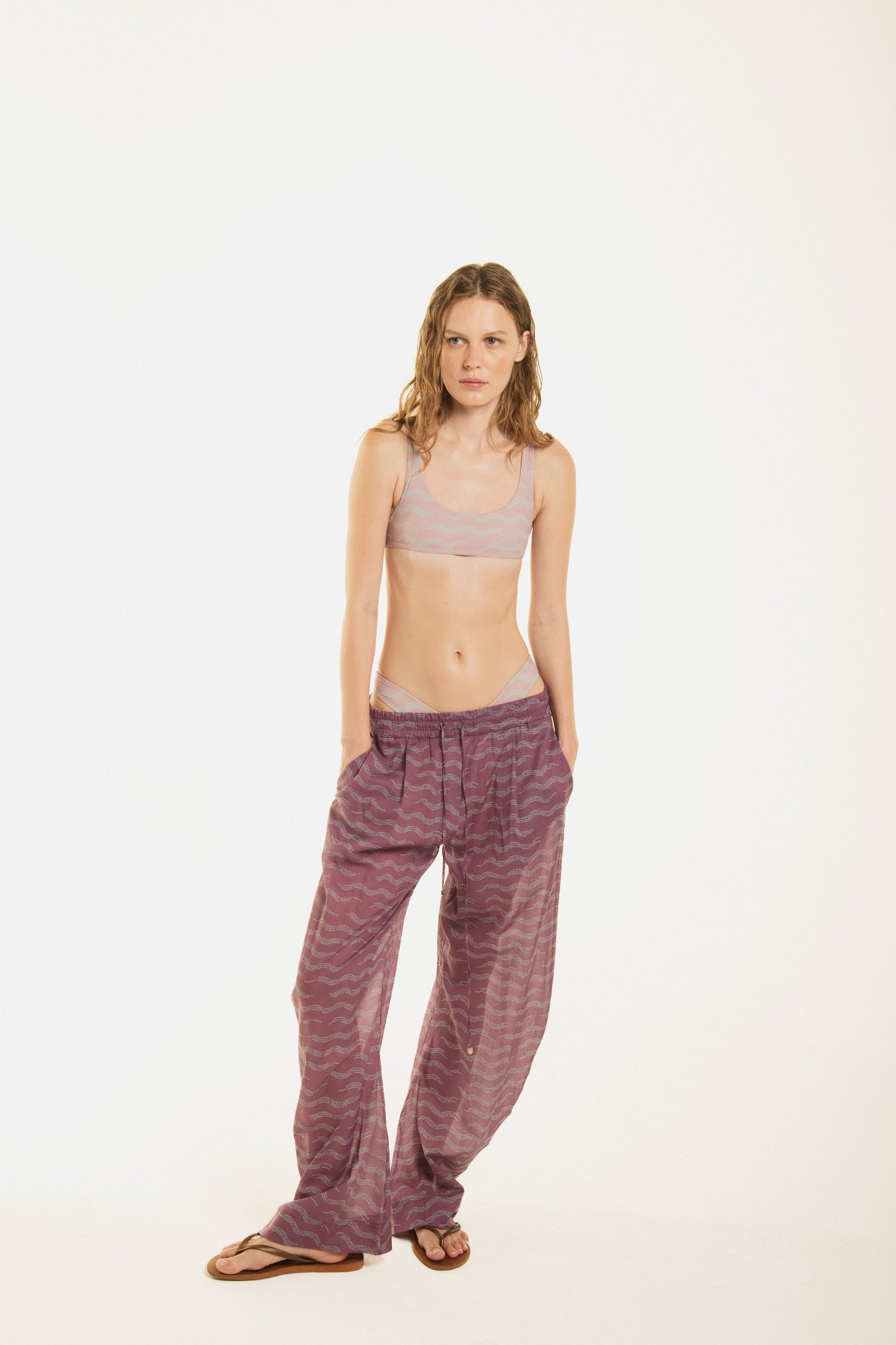 Front view of a girl wearing Nicasia grape violet muslin cotton lightweighted pants with an original print wave-inspired. Premium fabrics, oversized fit. The girl wears also Zoe pink bikini 